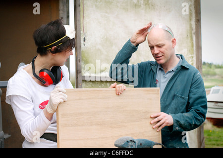 A man and a woman discussing the sanding work of a wooden door. Both are wearing Personal Protective Equipment (PPE) for safety. Stock Photo
