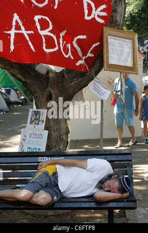 A tent city protester sleeping on a park bench under a banner "stop de abuse" hanged up in the Rothschild Boulevard in Tel Aviv. Stock Photo