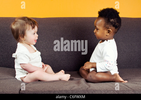 Two baby girls sitting on a sofa, looking at each other Stock Photo