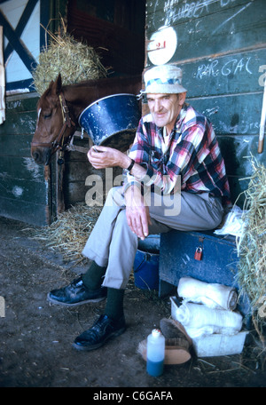 old groom poses for camera in front of horse stall and bandages Stock Photo