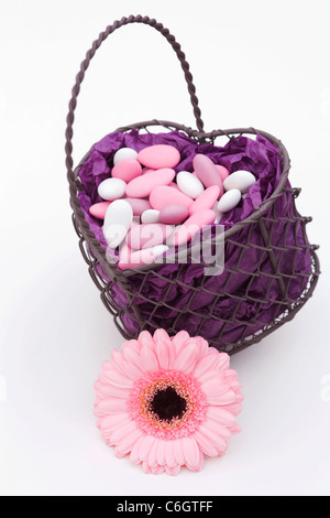 Sugared almonds in a heart shaped basket with a pink Gerbera on a white background. Stock Photo