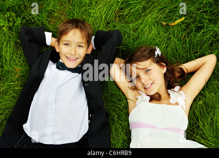 Portrait of smiling children bride and groom lying on green grass and looking at camera Stock Photo
