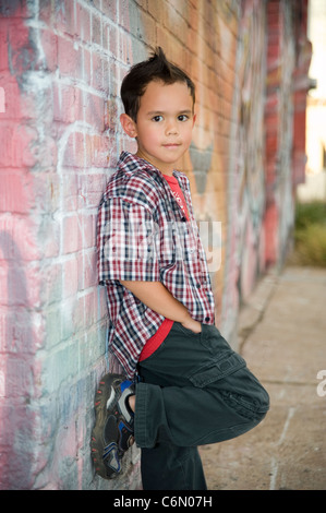 A cute 4-5 year old mixed race boy leans on a graffiti covered brick wall. Stock Photo