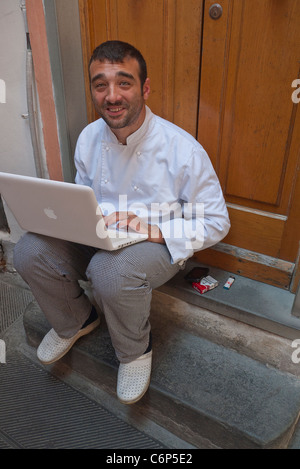 A young Italian male chef works on his laptop on the front step outside his restaurant in Riomaggiore, Cinque Terre, Italy. Stock Photo