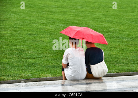 Two women sheltering under a red umbrella from the rain. View from the rear behind. Close up Stock Photo