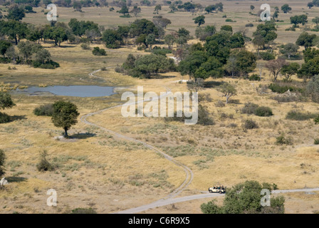 Aerial view over landscape with trees, dirt road and tourists on an open game drive vehicle approaching a waterhole Stock Photo