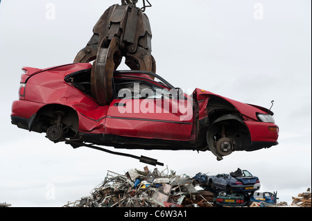 A red car is lifted by a crane in a scrap metal yard to  be crushed and recycled. Stock Photo