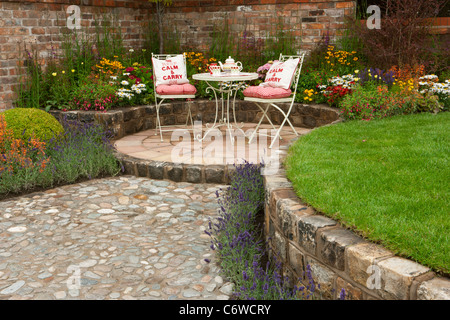 Small circle circular stone paved patio and lawn with a table and chairs seating with cobble cobbled pebble path and reclaimed brick raised bed beds Stock Photo