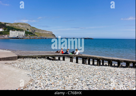 Young female students sun bathing on a wooden jetty on Llandudno seafront, background of sea, pier, great Orme with a blue sky. Stock Photo