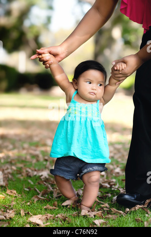 Baby girl taking her first steps with her mom helping her support her weight by holding her arms, walking on grass during fall Stock Photo