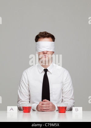 Studio portrait of young man with blindfold sitting in front of two glasses with red liquid Stock Photo