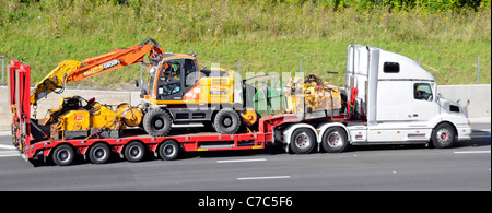 Side view white Volvo hgv bonneted traction unit and articulated low loader trailer loaded construction machines driving along UK motorway Stock Photo