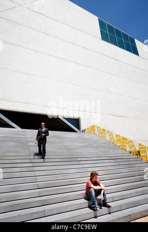 A guy who sits on the steps of the Casa da Musica in Porto is listening to music while a businessman is walking down the stairs Stock Photo