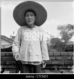 An historical photo from 1950s showing an eldery chinese worker posing for his picture wearing traditional hat and using cane. Stock Photo