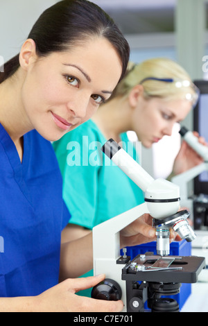 A beautiful female medical or scientific researcher using her microscope in a laboratory with her colleague behind her. Stock Photo