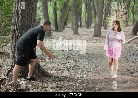 Overweight man hiding behind a tree, about to leap for young woman walking through the forest. Stock Photo