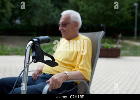 A senior man sitting in a chair, the handle of his walker in the foreground, focus on foreground Stock Photo