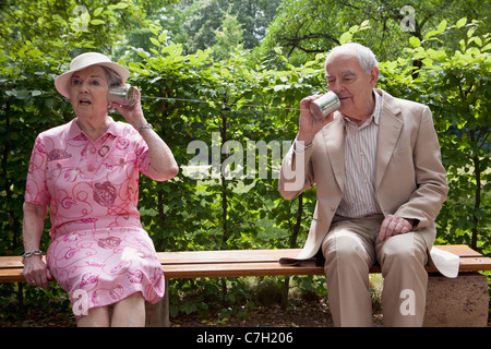 Senior man and woman speak to each other on tin can phones on bench in park Stock Photo