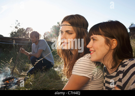 Two smiling girls in foreground with guy in background poking fire in field Stock Photo