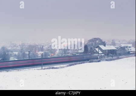 Snowy winter evening, passenger railway train speeding past rural village & white snow covered field - Wharfedale line, West Yorkshire, England, UK. Stock Photo
