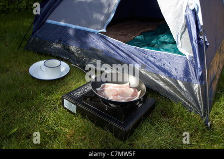 frying bacon on a small portable gas cooker in front of an open tent door Stock Photo