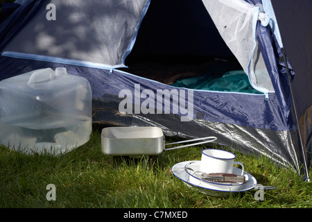 water container solid fuel stove and mess tin plate cup and camping tool utensils in front of tent open door Stock Photo