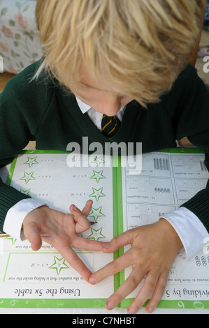 Blond-haired boy in green school uniform counts on his fingers doing his maths homework trying to add up sums sitting at the dining table Stock Photo
