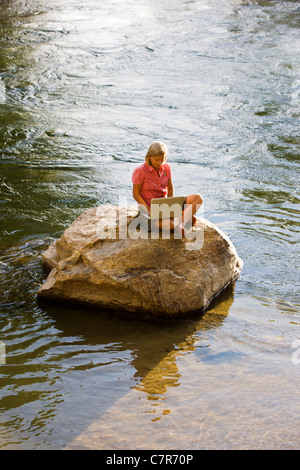 Lone woman sitting on a rock in a river working on a laptop computer Stock Photo