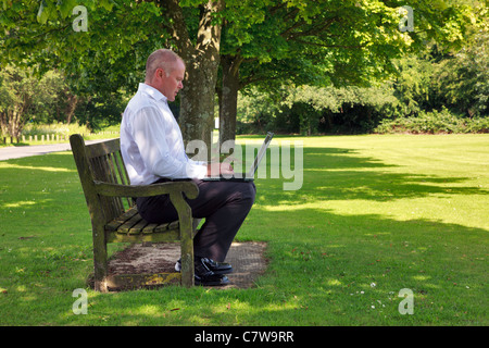 Photo of a businessman in smart casual clothing sat on a park bench working on his laptop computer. Stock Photo