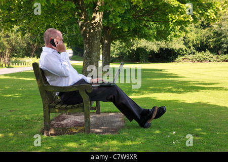 Photo of a businessman in smart casual clothing sat on a park bench working on his laptop computer. Stock Photo