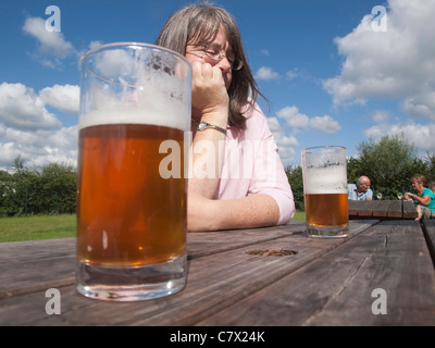 Thoughtful woman in a beer garden on a sunny day with half full glasses of beer on the plain wooden table Stock Photo