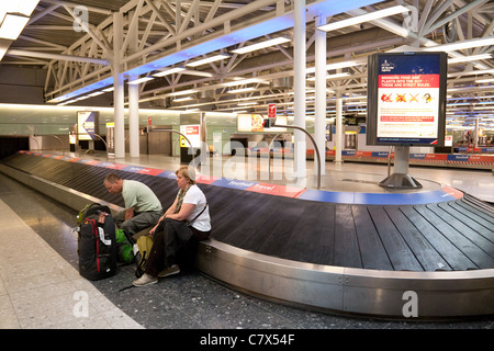 Lost luggage airport; 2 passengers sitting on the baggage carousel in the collection area having probably lost their luggage, Heathrow airport UK Stock Photo