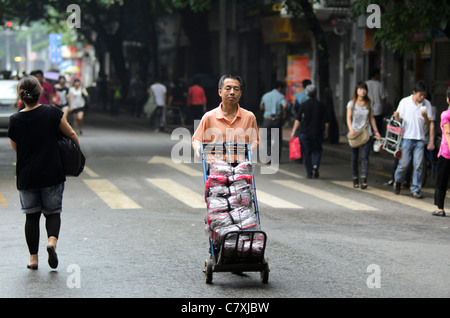 Chinese man (a worker) pushing a trolley down a the street in a city suburb, with people passing by. Stock Photo