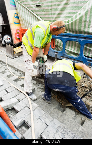London Camden Town workmen laying paving stones cobbles cobblestones with a pneumatic drill shovel spade high visibility hi vis jacket blue barrier Stock Photo