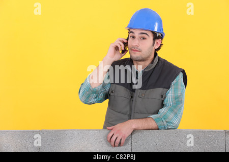 young bricklayer on the phone behind concrete wall against yellow background Stock Photo