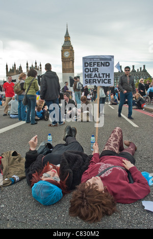 Two women lie down on Westminster Bridge with placard 'Defend our Health Service' behind is Big Ben. Health and Social Care Bill Stock Photo