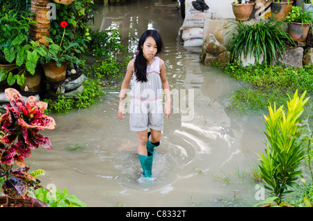 A cute 9 year old Asian girl with long black hair wearing boots walks through ankle deep water in a flooded yard in Thailand Stock Photo