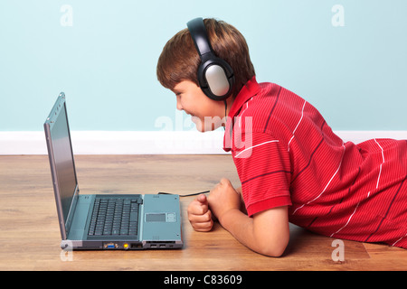 Photo of a boy lying on the floor wearing headphones and looking at the screen of his laptop Stock Photo