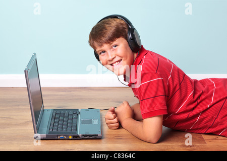 Photo of a boy wearing headphones looking towards camera lying on the floor next to his laptop computer. Stock Photo