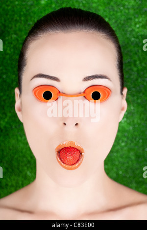 Closeup portrait of a woman wearing tanning bed glasses with strawberry in mouth against green Stock Photo