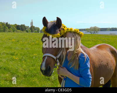 Young girl with her pony standing in a field in front of a lake. wreath of flowers on both the girls and the pony´s head. Stock Photo