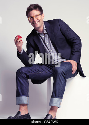 Fashionably dressed smiling man in his thirties wearing jeans and a stylish jacket holding an apple in his hand Stock Photo