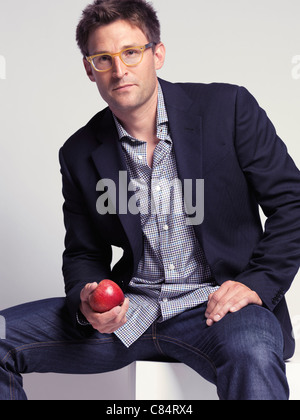 Fashionably dressed man in his thirties wearing jeans and a stylish jacket holding an apple in his hand Stock Photo