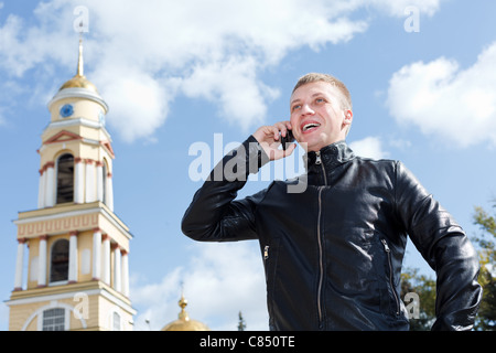 Young Russian handsome male on the mobile phone ringing outdoors near Church. Summer time, sunny day, blue sky. Russia Stock Photo