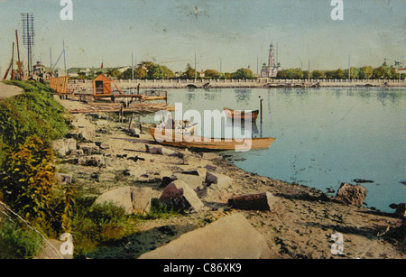 Boat station at the City Pond (Gorodskoy Pond) in Yekaterinburg, Russian Empire. Black and white vintage photograph by an unknown photographer dated from the beginning of the 20th century issued in the Russian vintage postcard published by Kh. T. Pvekov. The bell tower of the Great Zlatoust Church is seen in the background. The church was destroyed by the Bolsheviks in 1930s and restored in the beginning of the 21st century. Courtesy of the Azoor Postcard Collection. Stock Photo