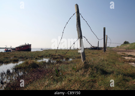 River Ore / Alde, bank / wall at Orford Suffolk, UK showing abandoned boats in background and barbed wire in foreground Stock Photo