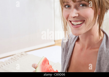 Young woman sitting on bed and holding watermelon slice in morning, smiling, close up, portrait