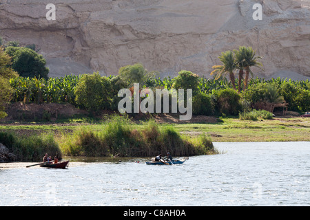 Section of lush green Nile river bank against a backdrop of rock with two small fishing boats on river in front, Egypt Stock Photo