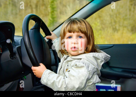 A toddler, a girl is at the steering wheel of a car playing at driving and looks into the camera, caught but also self-confident. Stock Photo