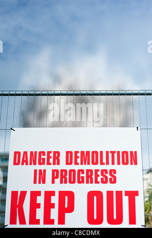 Sign attached to fence reading ' Danger Demolition in Progress KEEP OUT ' with rising smoke behind it. Stock Photo